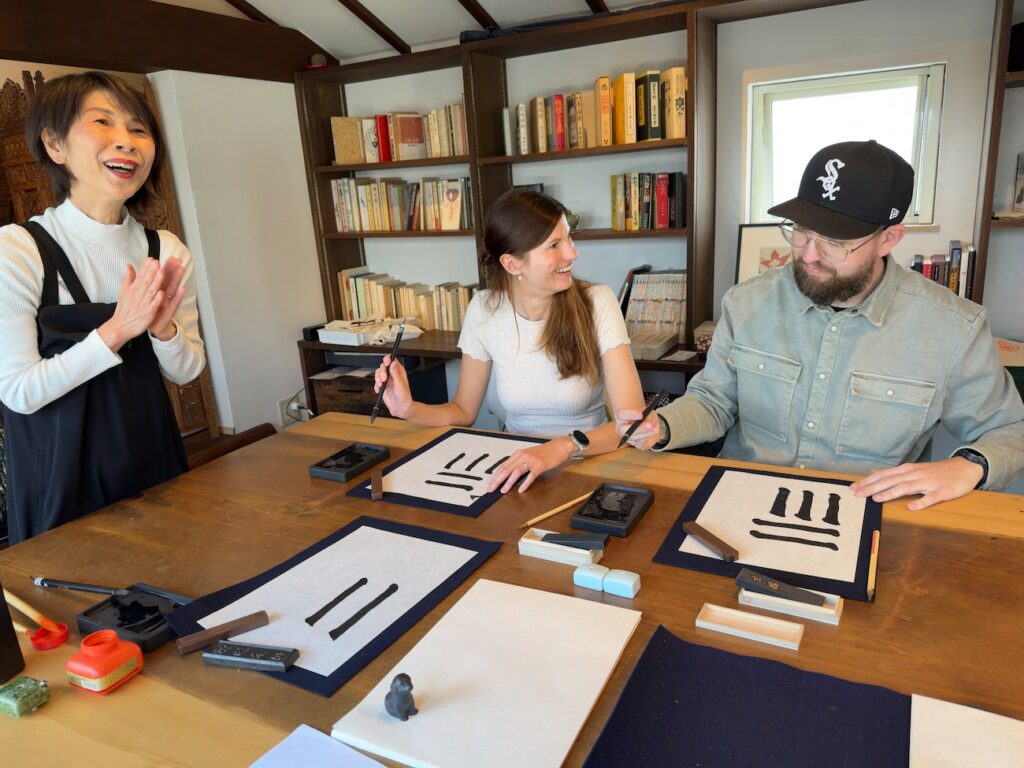 Two guests from Germany practicing basic calligraphy brush strokes with instructor guidance in Kamakura